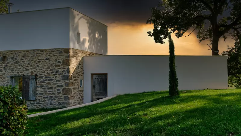 vue de la façade nord du gite la cubane dans le rouergue, gorges de l'aveyron en occitanie ; l'architecture est contemporaine ; on aperçoit la partie centrale sur deux niveaux et toit terrasse et l'aile ouest sur un niveau, la porte d'entrée en bois ancien, la pelouse et le soleil couchant