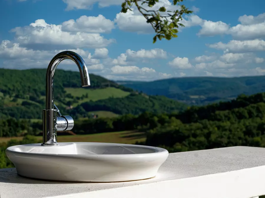 la cuisine extérieure du gite la cubane en occitanie ; évier vasque au premier plan avec le robinet chromé et en arrière plan, flou, le paysage formé de vallons boisés et de prés