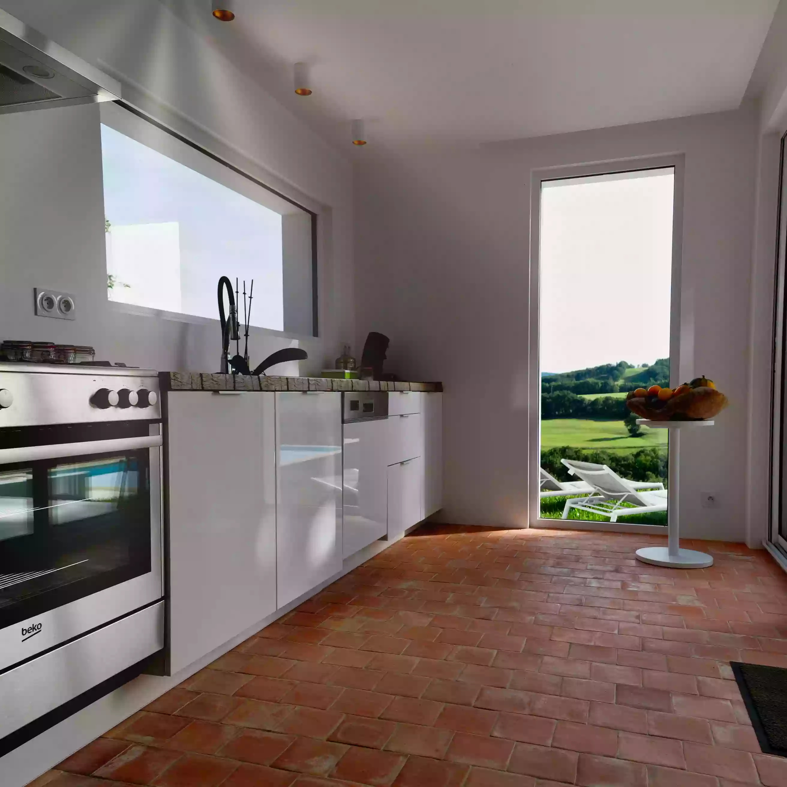 la cuisine équipée du gite la cubane à saint antonin noble val, gorges de l'aveyron ; sur la gauche on voit les éléments de la cuisine, le plan de travail en bois, une fenêtre rectangulaire au dessus du plan et dans le fond une ouverture qui permet de voir le paysage valloné ; également un table de bistrot avec une coupe de fruit dessus.