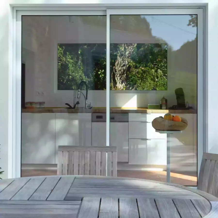 la cuisine du gite la cubane vue depuis la terrasse avec un jeu de reflet sur les baies vitrée ; une ouverture dans le mur du fond de la cuisine permet de percevoir les arbres en arrière plan