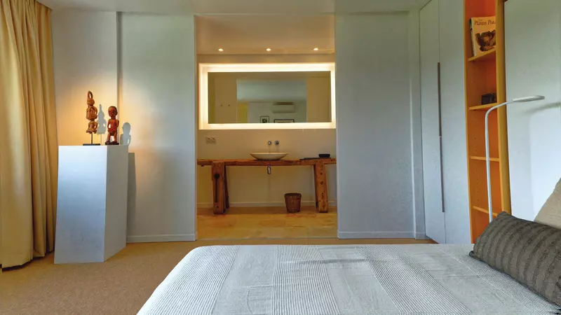 chambre du premier étage du gite la cubane en occitanie ; au premier plan, le lit, dans le coin gauche deux statuettes africaines sur un support rectangulaire, dans le fond la salle de bain avec sa vasque posée sur un établi réutilisé et sur le mur de droite un meuble intégré faisant bibliothèque ; éclairages d'ambiance