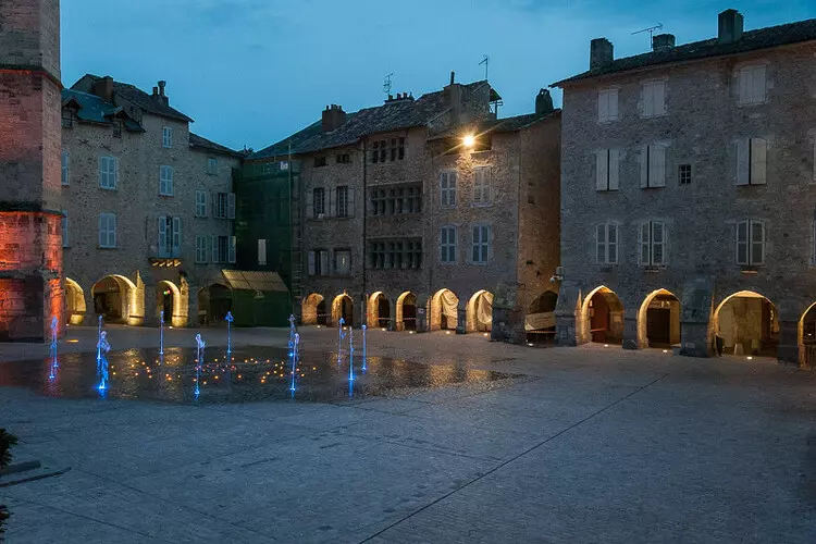 la place notre dame de villefranche de rouergue en aveyron, occitanie la nuit avec l'éclairage et la fontaine contemporaine en fonctionnement ; en rez de chaussée les arcades des maisons sont éclairées donnant une lumière d'ambiance